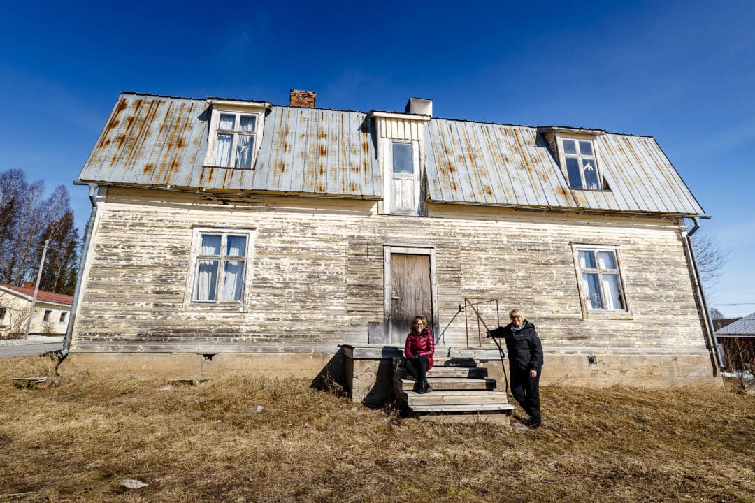 Två kvinnor framför ett gammalt stort hus med flagnad färg på väggarna. Blå himmel.